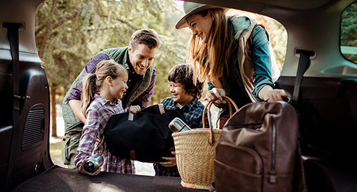 Family of four two kids two parents packing bags into the back of their van for vacation while talking and laughing