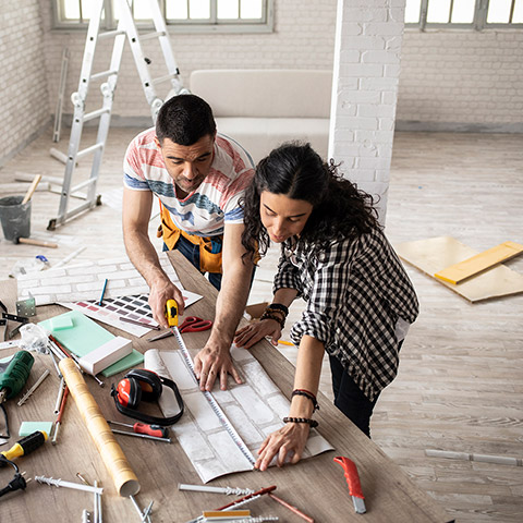 Middle age couple standing in a room that is being renovated the couple is looking at blueprints and measuring a table