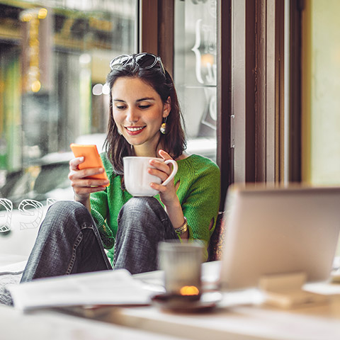 Woman sitting in a coffee shop looking at her mobile phone while holding a coffee mug